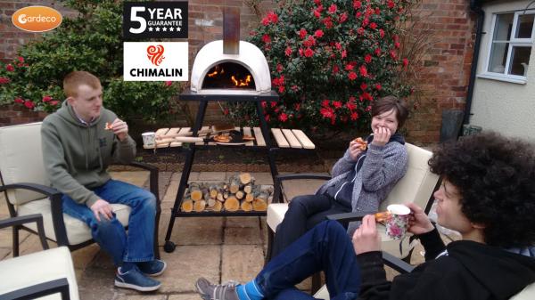 Three people enjoying pizza around a Chimalin outdoor pizza oven in a garden setting.