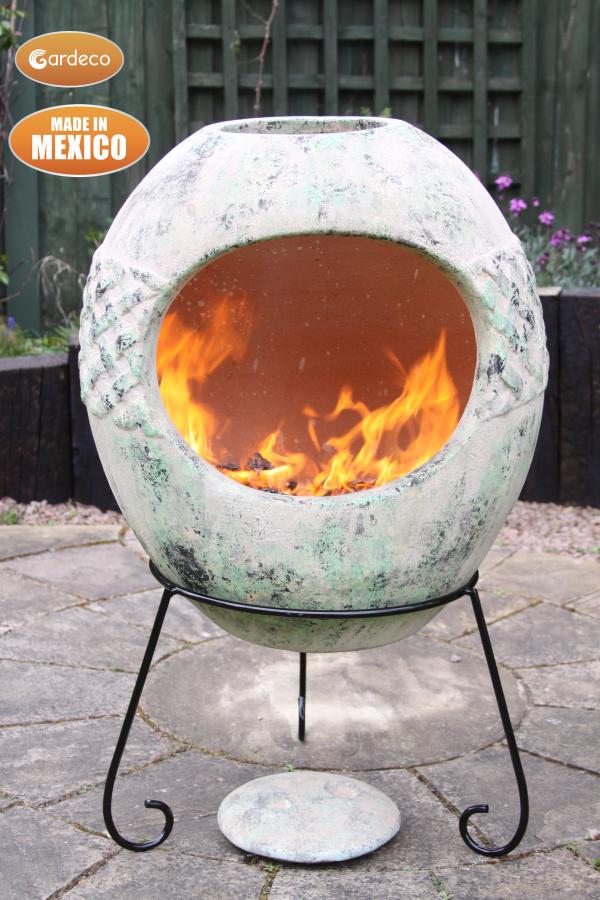 A spherical outdoor fire pit with a distressed green and white patina finish, shown lit with orange flames on a black metal stand in a garden patio setting. Gardeco and Made in Mexico badges are visible in the top left corner.